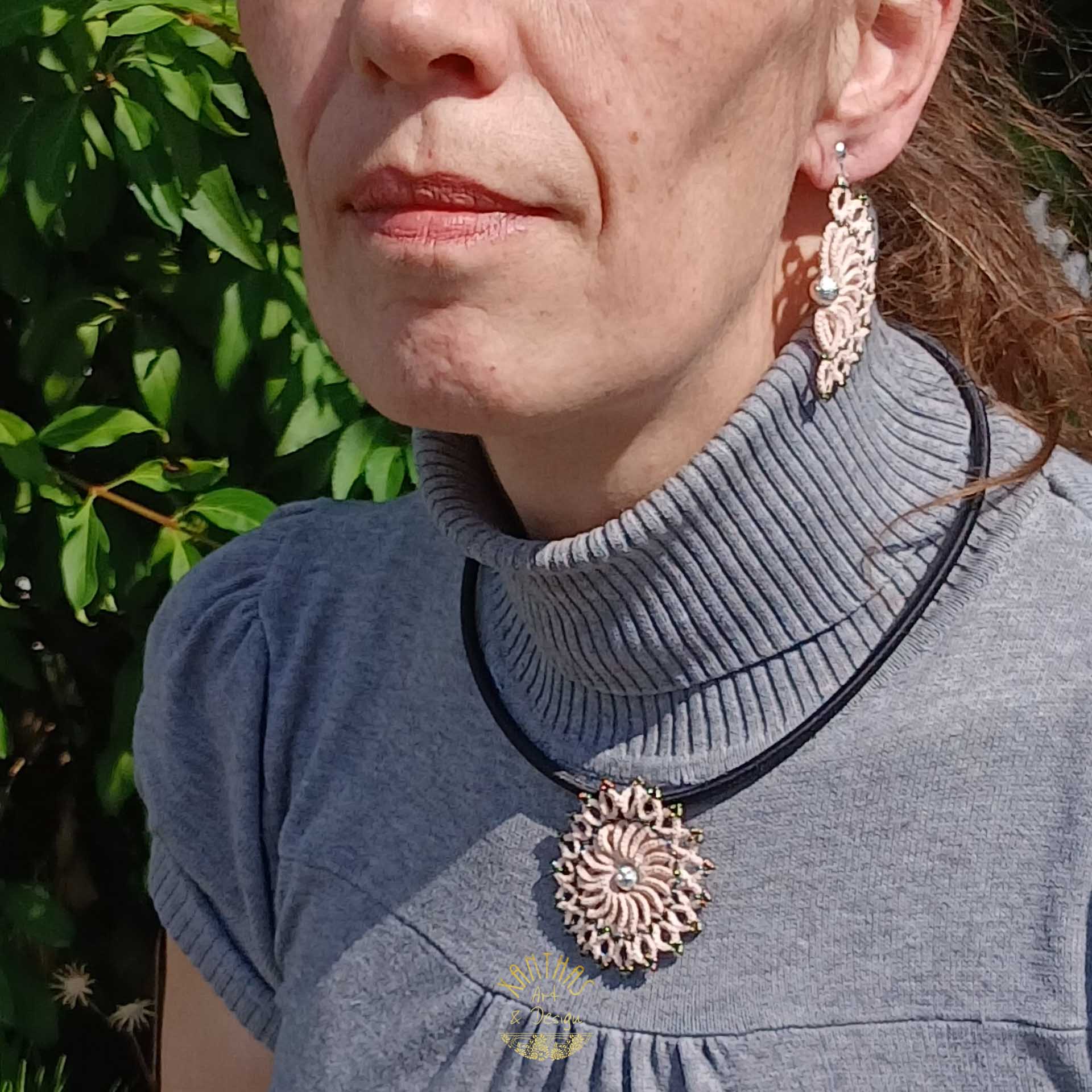Schmuckset „Clio“ aus handgefertigter Frivolitätenspitze in Zartrosa/Lachs. Das Collier hat einen runden Anhänger mit silber galvanisierter Lavaperle in der Mitte und bronzefarbigen Rocailles, kombiniert mit einer schwarzen Textilhalskette mit silberfarbenem Verschluss und Verlängerungskette. Die Ohrclips sind halbmondförmig mit silber galvanisierter Lavaperle und bronzefarbigen Rocailles, gefertigt aus 925 Sterling Silber.