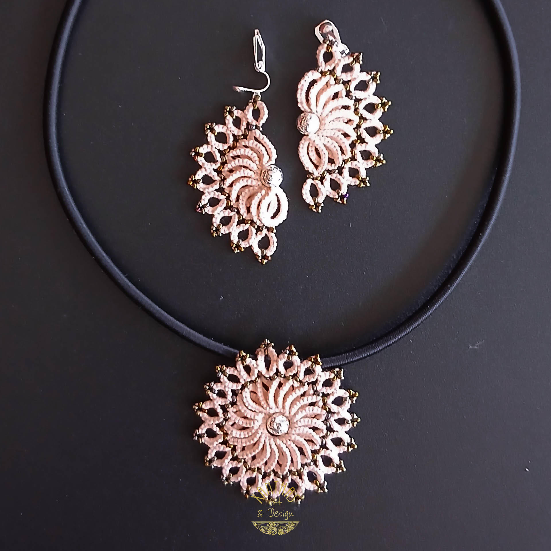 Schmuckset „Clio“ aus handgefertigter Frivolitätenspitze in Zartrosa/Lachs. Das Collier hat einen runden Anhänger mit silber galvanisierter Lavaperle in der Mitte und bronzefarbigen Rocailles, kombiniert mit einer schwarzen Textilhalskette mit silberfarbenem Verschluss und Verlängerungskette. Die Ohrclips sind halbmondförmig mit silber galvanisierter Lavaperle und bronzefarbigen Rocailles, gefertigt aus 925 Sterling Silber.